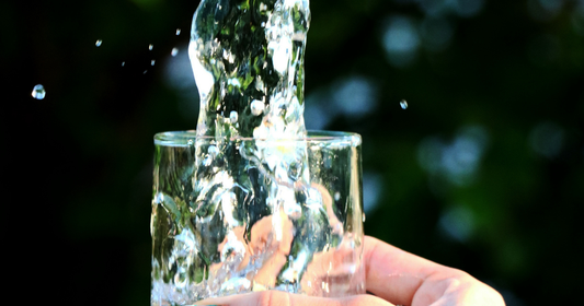 Kristallklart, rent vatten hälls upp i ett elegant glas, en visuell symbol för renhet, biologi och vikten av att filtrera sitt vatten hemma.