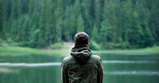 En man som står i total stillhet vid en spegelblank sjö omgiven av djup skog, en visuell symbol för mindfulness, tystnad och inre återhämtning i naturen.